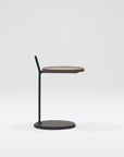 The Bystander Side Table by Wooden Stories features a round wooden top, slim black vertical support, and round black base, shown against a plain white background for a minimalist look.