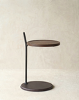 The Bystander Side Table by Wooden Stories features a compact, round solid wood top paired with a slender black metal rod and matching base, creating a modern accent piece for any space.