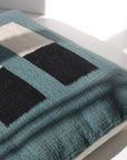 Pillow 04: Blue + Black by Ania Brzeski—a handwoven teal pillow with black and cream rectangles—rests in sunlight near a window, casting soft shadows on a white surface.