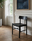 A SOBU Cesco Dining Chair in Black Ash is placed on a light wooden floor near a large window with white curtains. A potted plant sits nearby and a dark abstract painting hangs on the white paneled wall behind the chair.