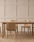 A minimalist dining room features the Wooden Stories Cove Dining Table in Light Oak, five matching wooden chairs, and a simple bowl, set against a beige panel wall with a neutral woven rug on the floor.