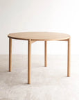 The Cove Dining Table | Light Oak by Wooden Stories, featuring a simple round design and four legs in solid American oak, stands on a textured floor against a plain white wall in a minimalistic setting.