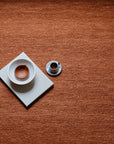 A small cup of espresso and a white bowl rest on stacked books atop the Armadillo&Co Cable Rug - Bisque, featuring Italian and Argentinian wools in a rich orange-brown hue.