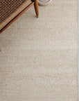 A woven wooden chair sits on the edge of an Armadillo&Co Cable Rug in Calico. A round, off-white ceramic object rests near the top right corner of the image.