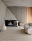 A minimalist living room with concrete walls, stacked firewood, a sculptural side table, and wood accents is grounded by the Armadillo&Co Cable Rug in Calico—a soft beige soumak weave—pairing beautifully with a cream sofa and modern wall lamp.