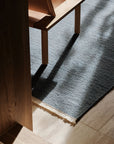 A wooden chair casts a shadow on the textured Cable Rug - Chambray by Armadillo&Co, its weave highlighted by sunlight streaming in and creating striking patterns on the light-colored floor.