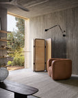 A cozy reading nook with a tan cushioned armchair, woven room divider, wall lamp, and the Armadillo&Co Cable Rug - Seed in soumak weave from Italian and Argentinian wools, set against concrete walls by a large window overlooking greenery.