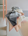 The Carte Postale Throw - Grey by Elvang Denmark, crafted from recycled wool in soft gray tones, is neatly folded and draped over the corner of a textured white cabinet, adding cozy elegance to a modern, minimal space.