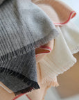 A close-up of the Elvang Denmark Carte Postale Throw - Grey, crafted from recycled wool in soft gray with subtle pinstripes and fringed edges. The fabric looks invitingly soft and cozy, accented with hints of cream, peach, and red.