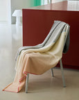The Carte Postale Throw - Grey by Elvang Denmark, featuring soft recycled wool in subtle tones, is draped over a white chair with a modern red table and minimalistic decor in the background.