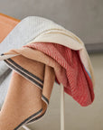 The Elvang Denmark Carte Postale Throw - Red Grey, featuring beige, gray, and red stripes, is folded and draped over a white metal chair set against a neutral background.