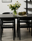 A modern dining room features the Wooden Stories Cove Dining Table in Black Oak, six black chairs, a flower vase, black shelves with decor, a wall mirror, a white armchair, a floor lamp, and minimalist art for an elevated dining experience.