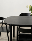 A minimalist dining area features the Wooden Stories Cove Dining Table in Black Oak, three black chairs, a glass vase with white and yellow flowers, and a framed hand photo on the white wall—perfect for an elevated dining experience.