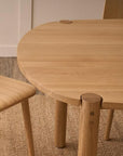 A light wooden chair and the Cove Dining Table - Light Oak by Wooden Stories sit on a woven rug atop a wooden floor, set against a white wall. The simple, modern design creates an inviting dining atmosphere.