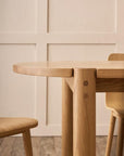 Close-up of the Wooden Stories Cove Dining Table in Light Oak paired with chairs, set against a beige paneled wall and woven rug on wood floor, creating a minimalist, cozy vibe inspired by relaxed Sydney Harbor dining.