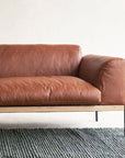 The Department Modular Sofa | Leather • M6 by Wooden Stories, featuring a modern brown leather finish and solid wood base, is placed on a textured gray rug against a white wall with only its left side visible.