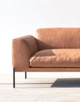 The Wooden Stories Department Modular Sofa | Leather • M5 Right features modern tan leather upholstery, slim black metal legs, and a solid wood base. Sunlight highlights its left side and seat cushion against a plain white wall on a light floor.