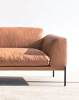 The Department Modular Sofa | Leather • M6 by Wooden Stories, featuring a minimalist tan leather design with a solid wood base and black metal legs, sits in sunlight against a white wall, casting a soft shadow on the floor.
