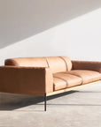 The Department Sofa 93" by Wooden Stories, in tan leather with a solid wood base and black metal legs, sits in a sunlit minimal room—its sleek silhouette casting bold shadows on the white wall and gray floor.