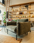 A modern living space featuring the Attendant Lounge Chair by Wooden Stories with black legs, a tan rug, and wooden shelves holding decor, plants, and tableware. A tall ladder leans against the wall in the background.
