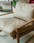 The Troubadour Lounge Chair | Leather • Light Oak by Wooden Stories features a modern Danish design with tan leather cushions and a light oak frame, placed on a tiled floor in a stylish, well-lit room with plants and shelves in the background.