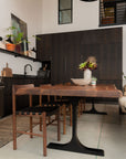 Modern kitchen and dining area with dark wood cabinets, wooden chairs, minimalist decor, a potted plant centerpiece, and the SOBU Leviathan Live Edge Dining Table in smoked acacia with black T-legs bathed in natural overhead light.