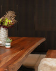 The SOBU Leviathan Live Edge Dining Table in smoked acacia with brass T-legs holds a white vase of green and burgundy flowers and a small succulent. A furry cushion is on a wooden bench nearby, with dark cabinets behind.