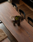 The SOBU Leviathan Live Edge Dining Table in smoked acacia with brass T-legs is paired with black chairs, a matching bench with a fur throw, decorative plants, a wooden bowl, a patterned rug, and a stainless steel oven nearby.