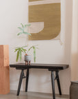 A modern, minimalist space featuring the SOBU Mars Live Edge Bench in Black Ash with natural wood details, topped with a potted plant and accented by a beige and green geometric wall hanging above, plus neutral walls and flooring.