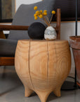 The SOBU Saturn Side Table - Natural, made of solid pine with a matte lacquer finish, features three legs. A vase of yellow billy balls and a black sphere sit on top, with a gray armchair in the background.