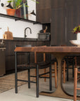 A modern kitchen with dark cabinets, the SOBU Leviathan Live Edge Dining Table with Brass T-Legs, black chairs, a countertop microwave, potted plants, a patterned rug, and shelving with decor above the sink.