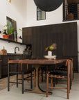 Modern kitchen and dining area with dark wood cabinets, plants, wall art, woven black and brown chairs, a large round black pendant light, and the SOBU Leviathan Live Edge Dining Table in smoked acacia with brass T-legs.