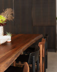A close-up of the SOBU Leviathan Live Edge Dining Table in smoked acacia with brass T-legs, surrounded by black and brown chairs. A white potted plant sits at one end, set against a dark, blurred background.