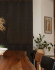 A close-up of SOBUs Leviathan Live Edge Dining Table in smoked acacia with brass T-legs, styled with a centerpiece bowl and chairs in a modern interior featuring dark cabinets, a potted plant, and framed art on a white wall.