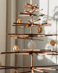The Immortal Christmas Tree by SOBU is a minimalist, sustainable piece crafted from horizontal branches and decorated with simple white and gold ornaments. Sunlight creates soft shadows on the wall behind it.
