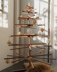 The SOBU Immortal Christmas Tree with geometric branches stands by a sunlit window, adorned with gold and white ornaments. A cozy brown fur rug rests at its base, creating a minimalist and sustainable holiday scene.