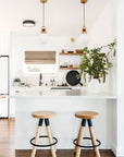 Modern kitchen with white walls, wooden floor, two SOBU Blix Counter Stools at a white island with modern steel frame, pendant lights, open shelves, potted plant, and neatly arranged kitchen items from the Blix Collection.