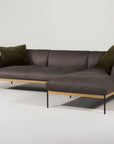 The Wooden Stories Department Modular Sofa | Leather • M2 Right features dark gray leather, a solid wood base, slim black metal legs, and two olive green pillows, displayed against a plain white background.