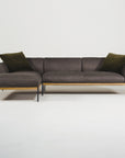 The Wooden Stories Department Modular Sofa | Leather • M2 features dark grey upholstery, a solid wood base, slim black legs, a left-side chaise lounge, and two dark green pillows, all set against a plain white background.