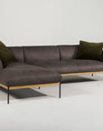 The Department Modular Sofa | Leather • M2 by Wooden Stories showcases dark gray leather, an FSC-certified oak base, black metal legs, and two olive green pillows, set against a plain white background.
