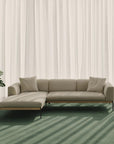 A beige Department Modular Sofa | Fabric • M2 by Wooden Stories with three cushions rests on a green carpet before sheer white curtains. Sunlight casts diagonal shadows, and a leafy green plant appears on the left side of the scene.