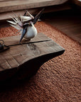 A rustic wooden coffee table rests on the Mason Rug - Sangria by Armadillo&Co, crafted from New Zealand wool. A white vase with spiky dried branches sits atop, while natural light enhances the cozy, minimalist ambiance.