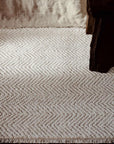 A close-up of the Armadillo&Co Crest Rug in Poplin, featuring a cream-colored herringbone pattern, displayed next to dark wood furniture and a beige fabric backdrop.