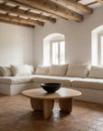 A minimalist living room with white walls, exposed wooden beams, a large white sectional sofa, terracotta tile flooring, and the Eden Coffee Table by Wooden Stories. Sunlight streams through arched windows, highlighting Scandinavian simplicity.