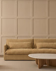 A beige textured sofa with large cushions faces a paneled beige wall, next to the Wooden Stories Eden Coffee Table | Wood & Stone, featuring a round oak base and white top, all set on a light rug in a minimalist Scandinavian-inspired space.