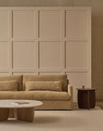 A minimalist living room inspired by Scandinavian simplicity, featuring a beige sofa, two matching cushions, a Wooden Stories Eden Coffee Table | Wood & Stone with a book, a dark side table with a cup, and a paneled light wall in the background.