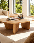 A modern living room with the Eden Coffee Table by Wooden Stories, adorned with books and a sculpture, is surrounded by beige sofas and large windows framing a lush garden—capturing true Scandinavian simplicity.