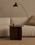 A minimalist living room with a beige sofa, textured cushion, the Wooden Stories Eden Side Table holding a small cup, a beige rug, and a black wall-mounted lamp against a light brick wall embodies Scandinavian simplicity.