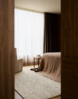 A cozy bedroom with a large window and sheer white curtains, brown bedding, a beige armchair, a side table with a cup, and an Armadillo&Co Faroe Rug - Foal in chunky weave on the wooden floor.