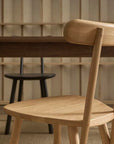 A Figura Dining Chair in Light Oak by Wooden Stories sits before a dark table, with two darker chairs partially visible against a heritage wood-paneled wall.